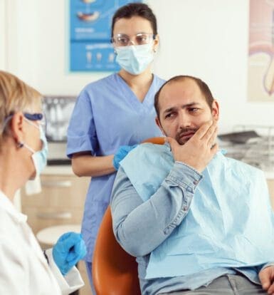 A male patient holding his jaw and looking concerned while talking to a female dentist and a dental assistant in a clinic.