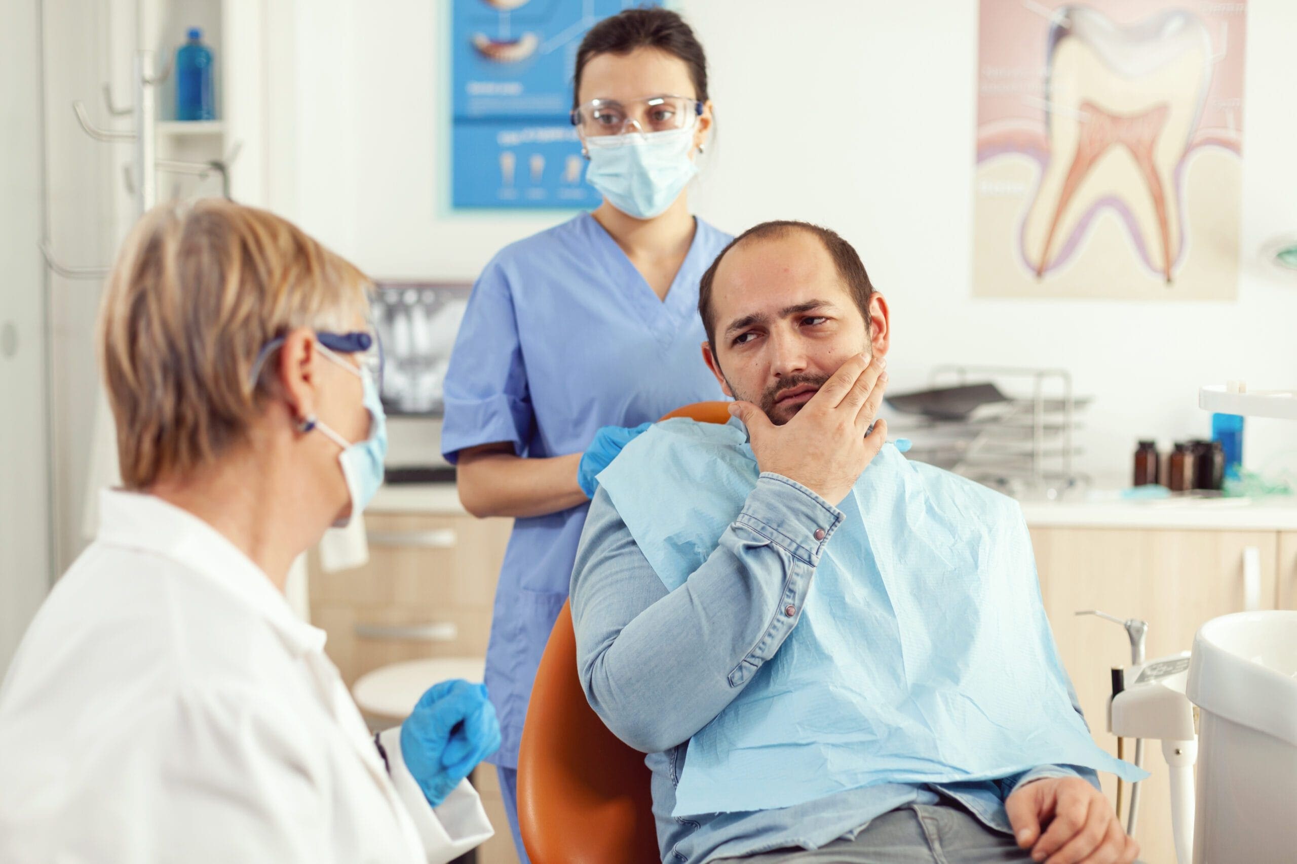 A male patient holding his jaw and looking concerned while talking to a female dentist and a dental assistant in a clinic.