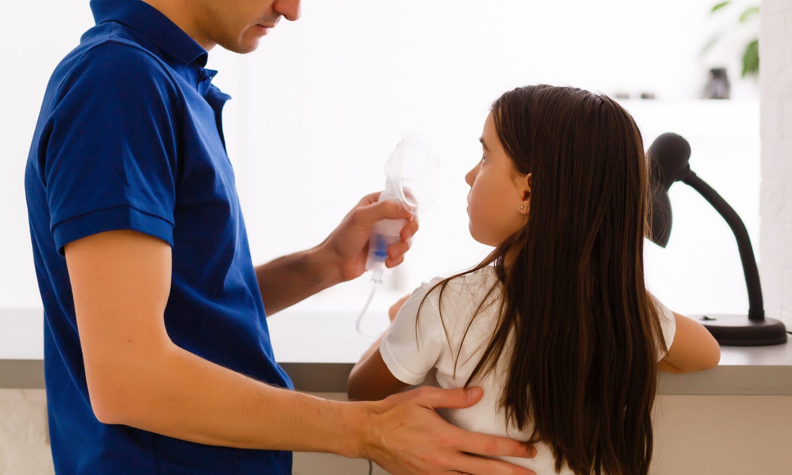 A young girl receiving a respiratory treatment with a nebulizer, assisted by an adult wearing a blue shirt.