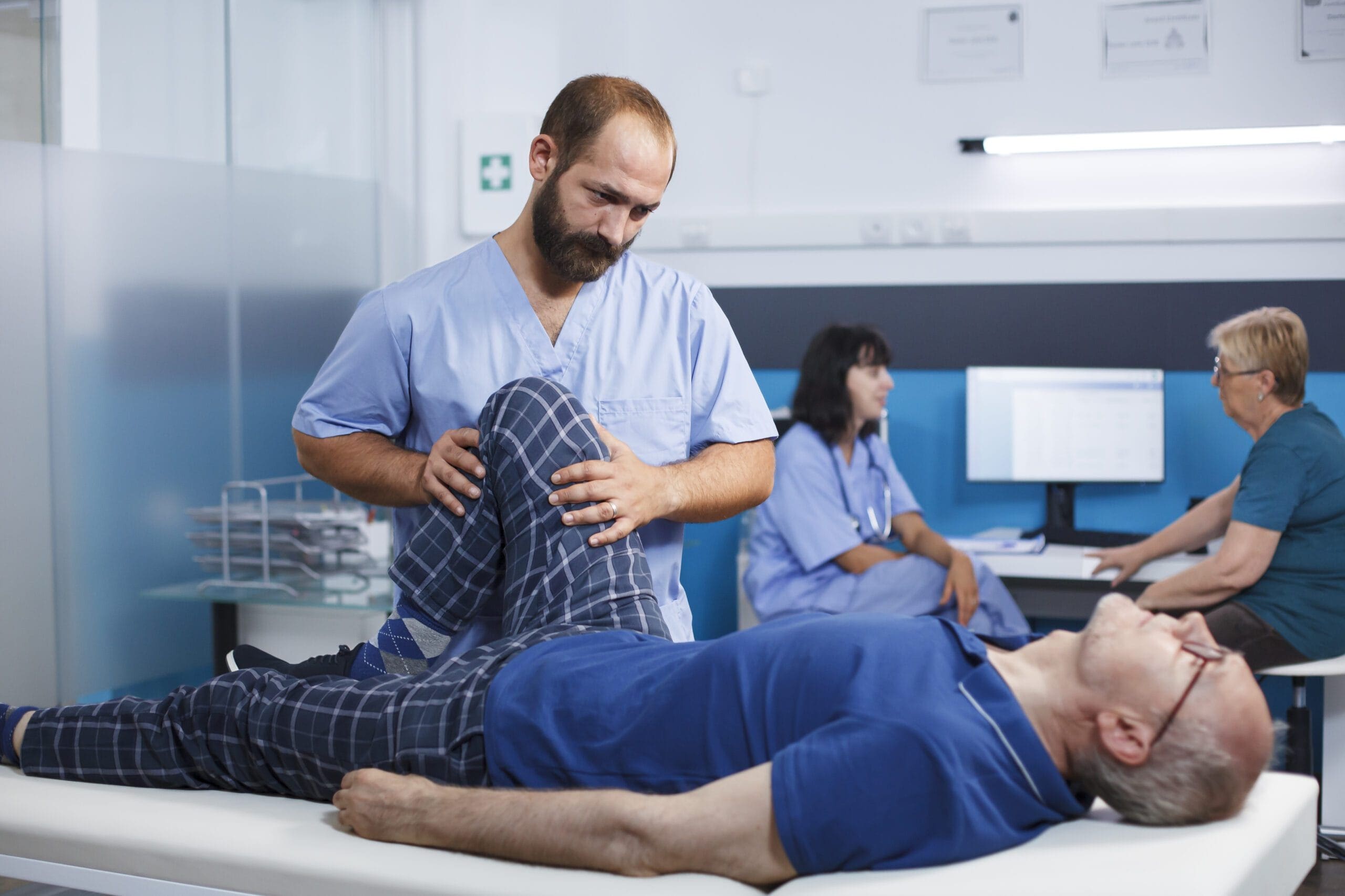 A healthcare professional examining an elderly patient's knee, with other medical staff in the background.