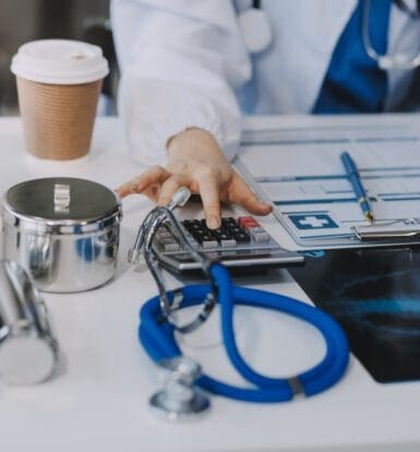 A healthcare professional using a calculator at a desk with medical tools, documents, and a stethoscope, representing medical coding and compliance.