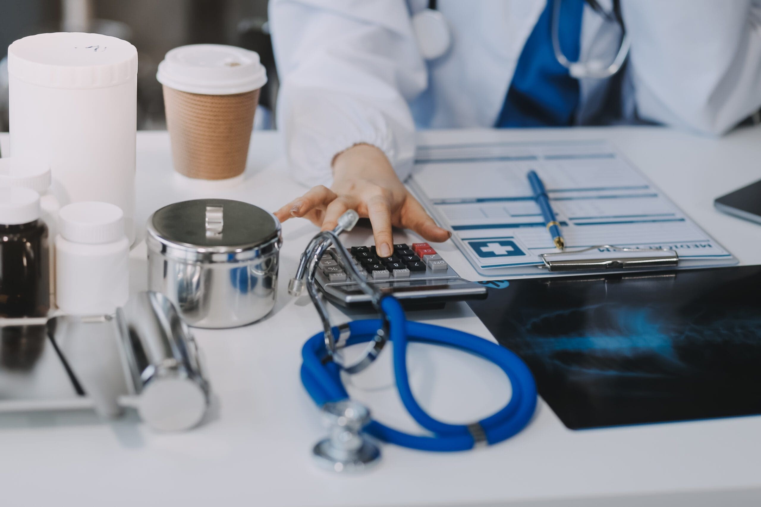 A healthcare professional using a calculator at a desk with medical tools, documents, and a stethoscope, representing medical coding and compliance.