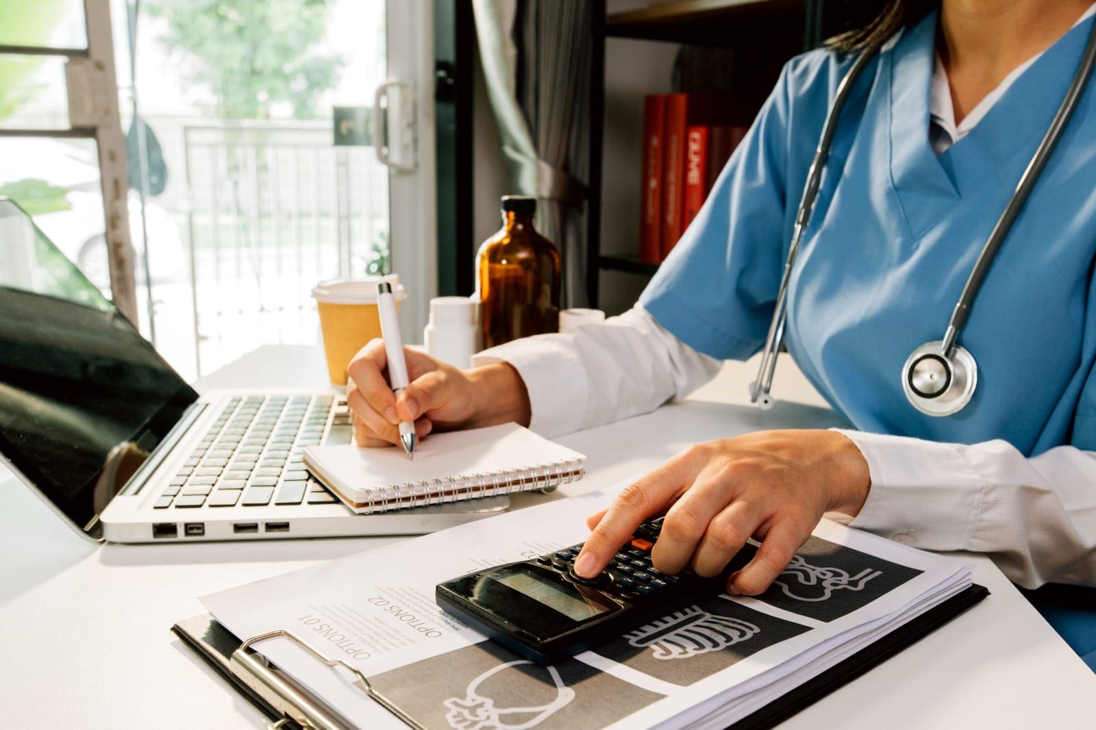 A healthcare professional in scrubs working at a desk, writing in a notebook while using a calculator, with a laptop and medical documents in the background.