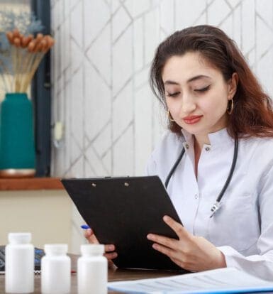 A female healthcare professional in a white coat reviewing a clipboard, sitting at a desk with medication bottles and documents in front of her