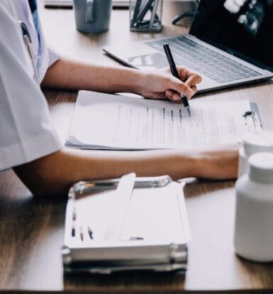 A healthcare professional reviewing medical forms and documentation at a desk, with a laptop and medication bottles nearby.