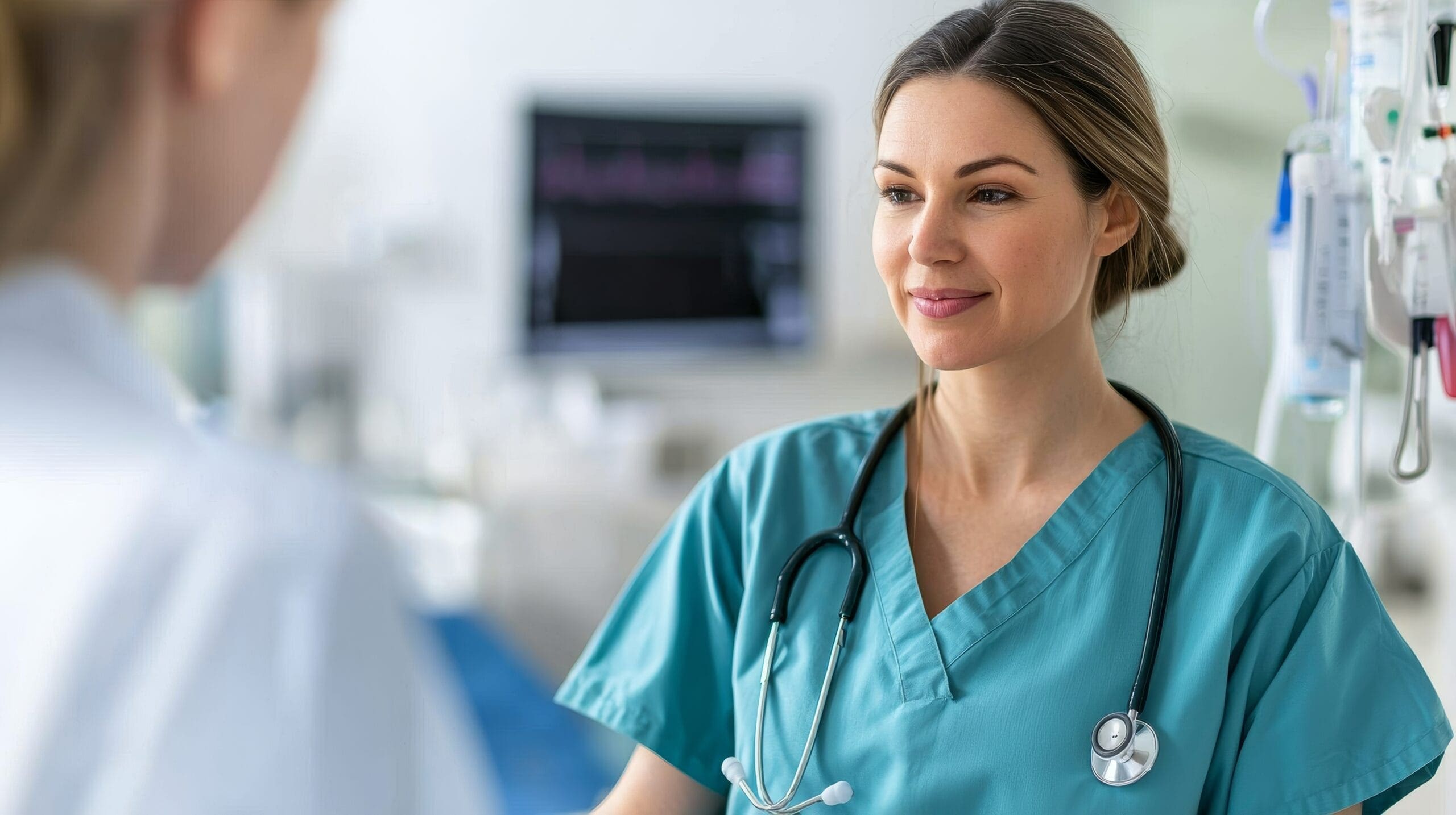 A smiling healthcare professional in teal scrubs with a stethoscope around her neck, standing in a medical facility