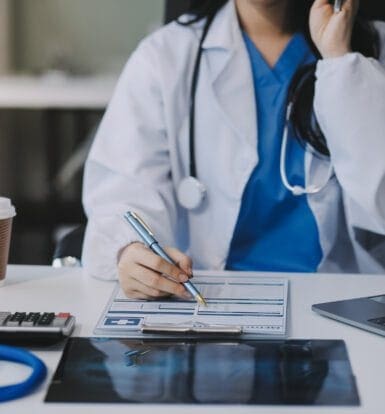 A healthcare provider in a rural clinic filling out medical forms with a stethoscope, laptop, and X-ray on the desk, symbolizing the importance of accurate medical coding for effective reimbursement and patient care.