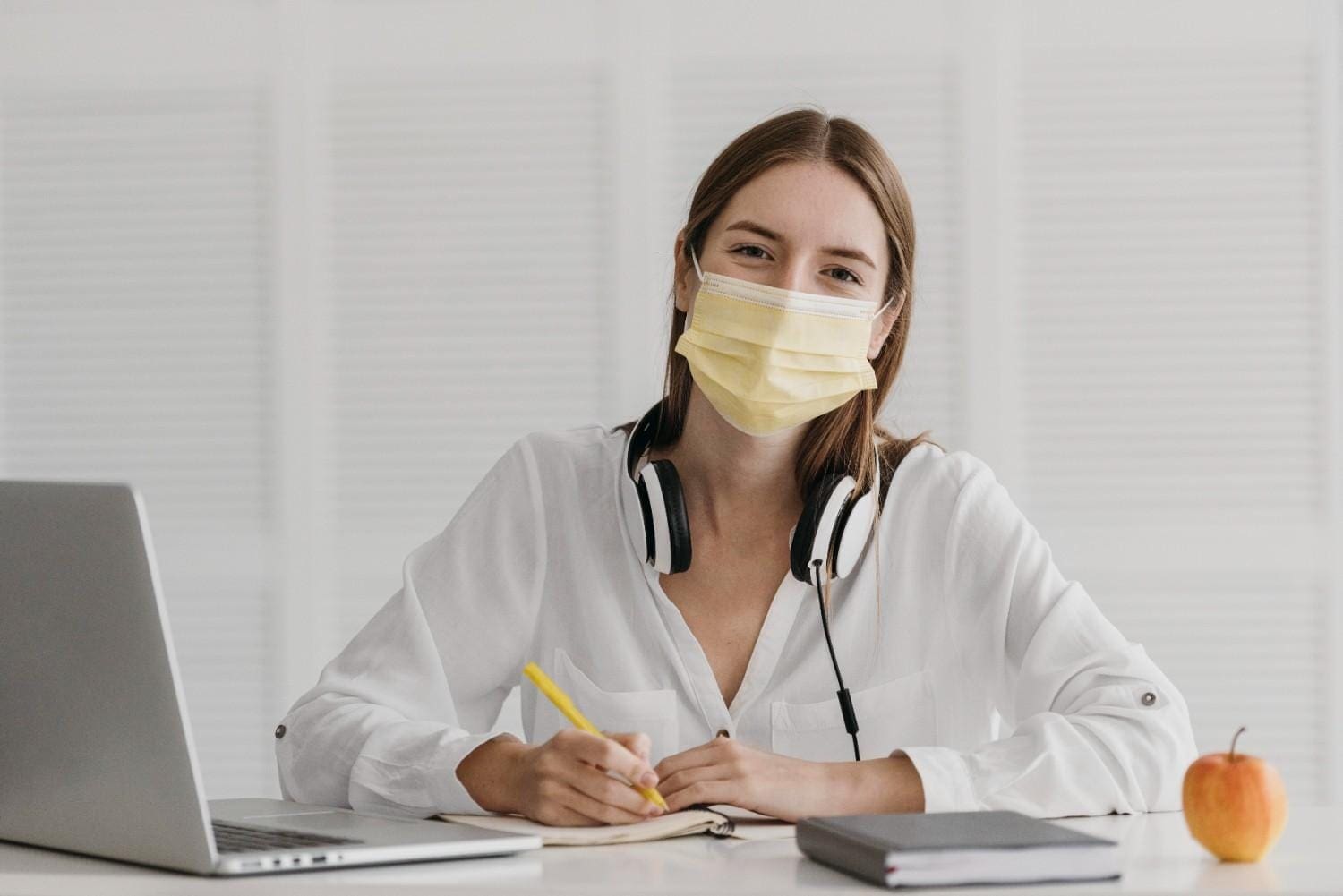 Young woman working remotely with headphones, mask, and laptop at a desk.