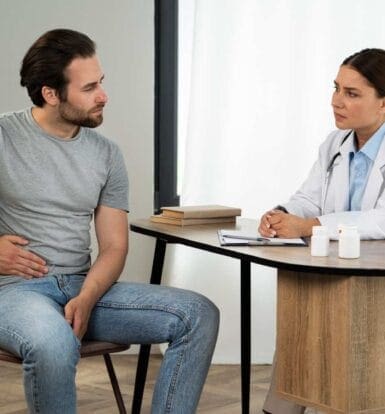 A concerned male patient holding his abdomen, sitting across from a doctor who is attentively listening, symbolizing a consultation about Xifaxan prior authorization for IBS-D or hepatic encephalopathy.