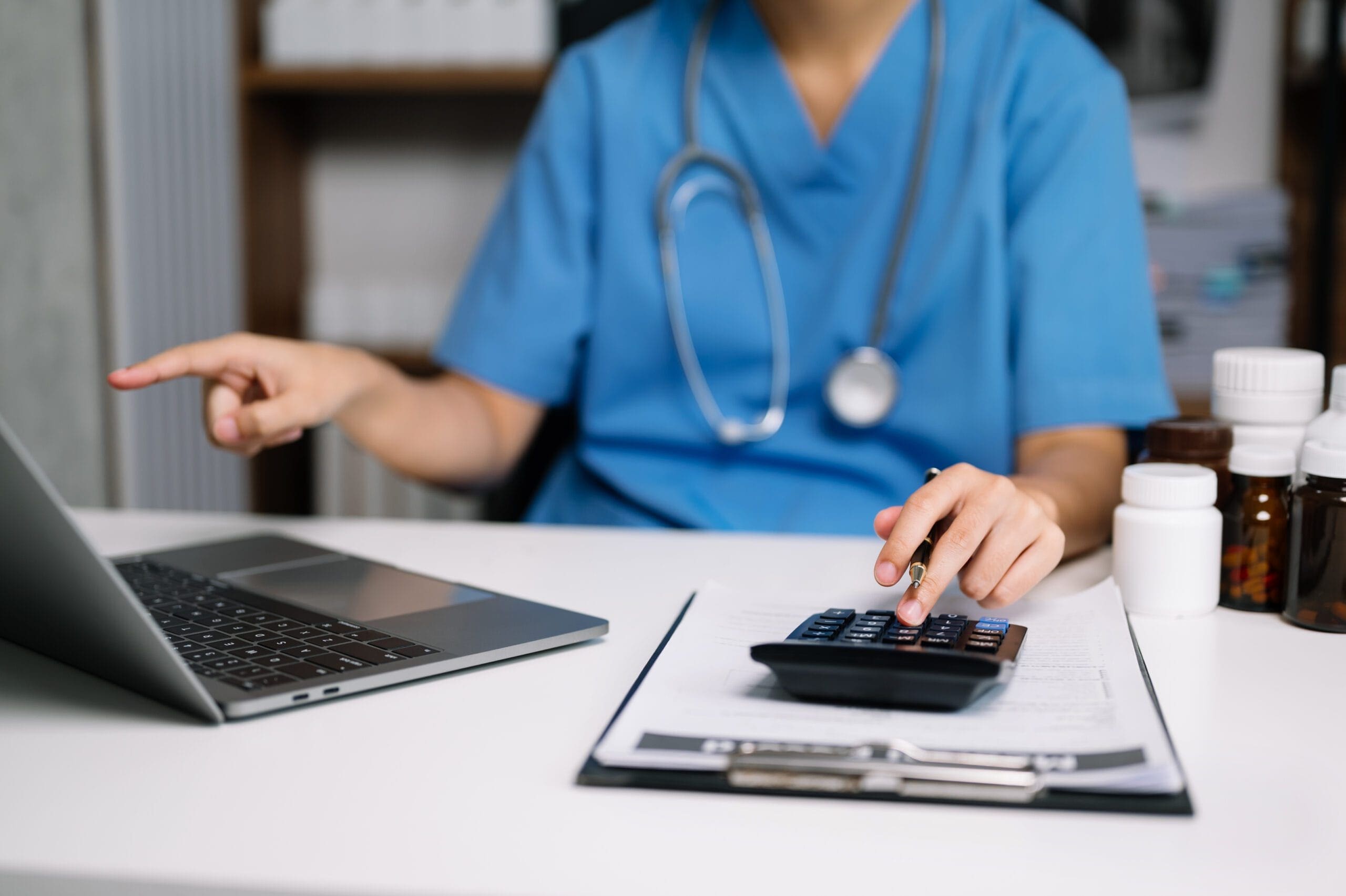 A healthcare professional in scrubs using a laptop and calculator with medication bottles on the desk, symbolizing the administrative and clinical coordination required for specialty pharmacy services.