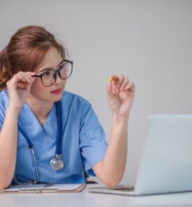 A nurse in blue scrubs sitting at a desk, examining a pill closely while working on a laptop, with bottles of medication nearby.