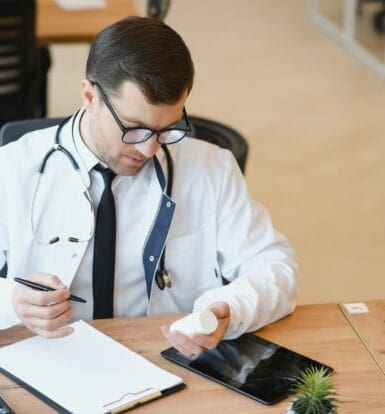 A doctor reviewing medication details while taking notes on a clipboard, with a tablet and phone on the desk.