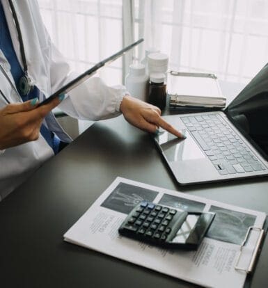 A healthcare provider using a tablet and laptop to manage EHR documentation, alongside medical tools and records on the desk.