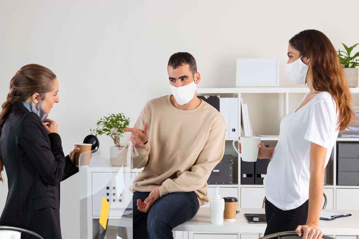 Three colleagues wearing masks discussing in an office environment, holding coffee mugs, with hand sanitizer on the table.