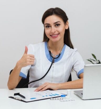 A smiling virtual medical assistant giving a thumbs-up while working at a desk with a laptop and healthcare documents.