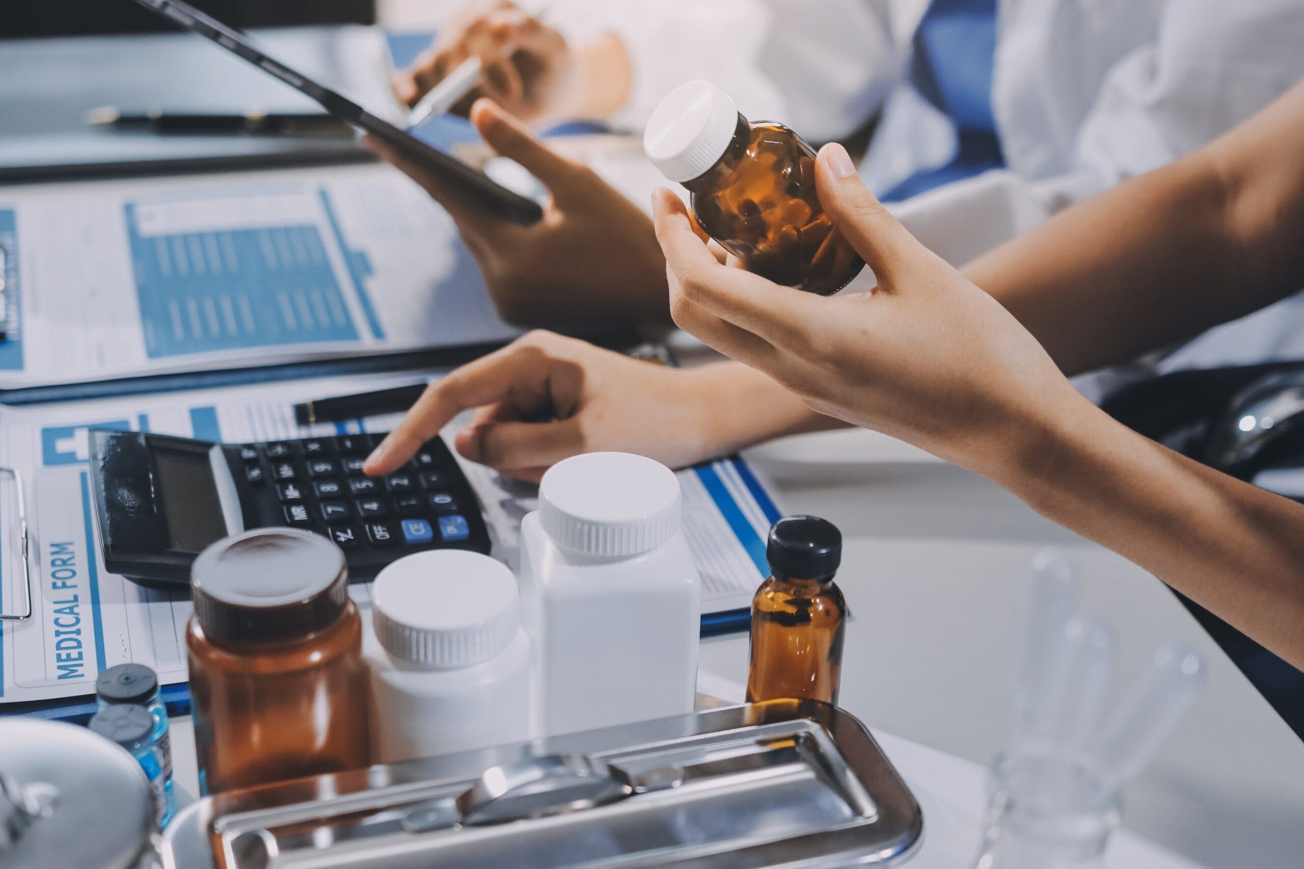 Close-up of healthcare professionals handling pharmacy billing tasks, with one person holding a medication bottle and another working on a calculator. The image includes medical forms, pill bottles, and a tablet, illustrating the complexity of LTC pharmacy billing processes.