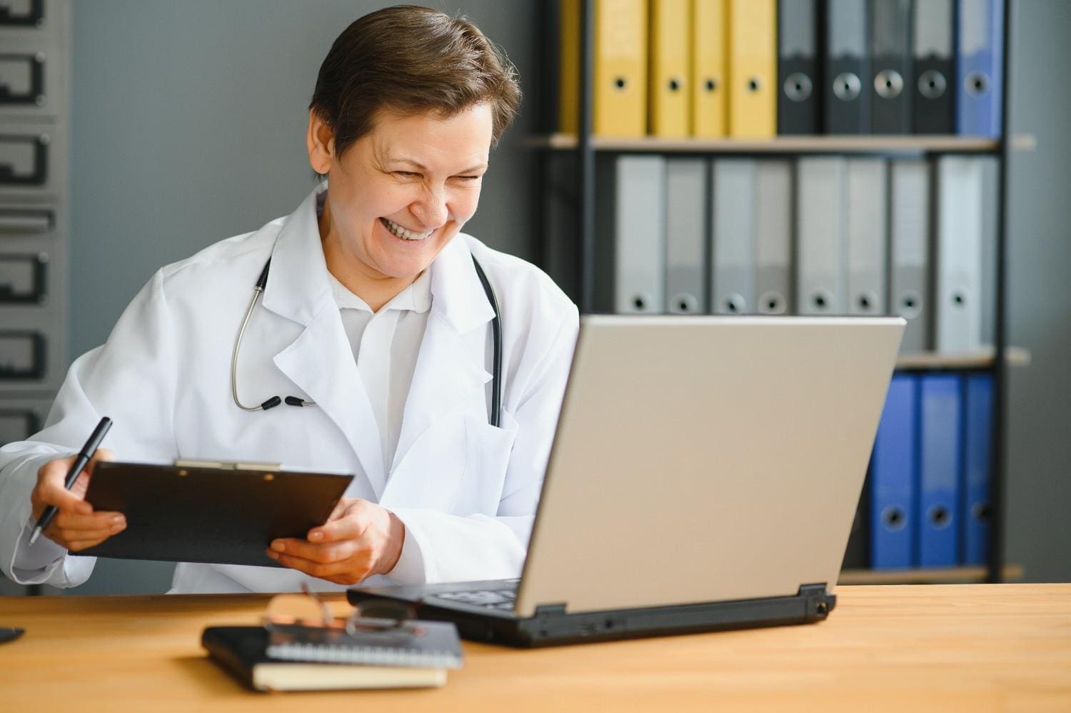Happy doctor using a laptop while taking notes, representing the efficiency of Virtual Medical Receptionist Services.